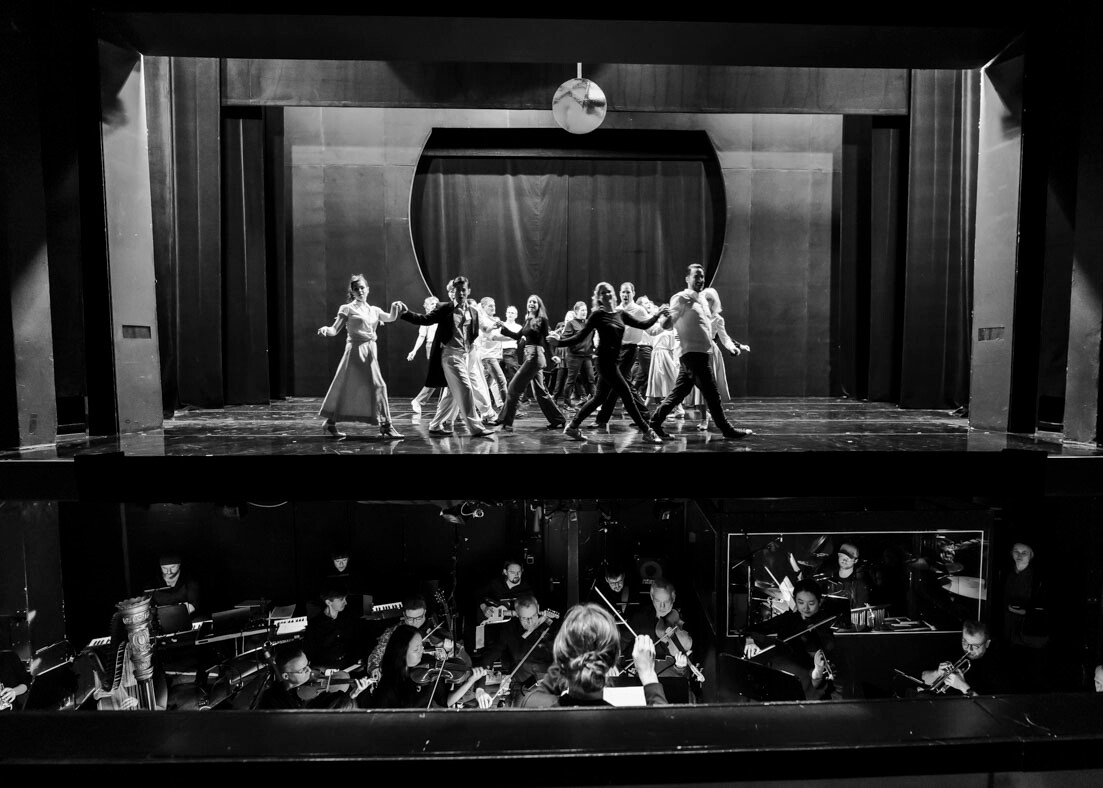 Ensemble tanzt auf der Bühne des Theaters Lüneburg, während das Orchester im Orchestergraben spielt