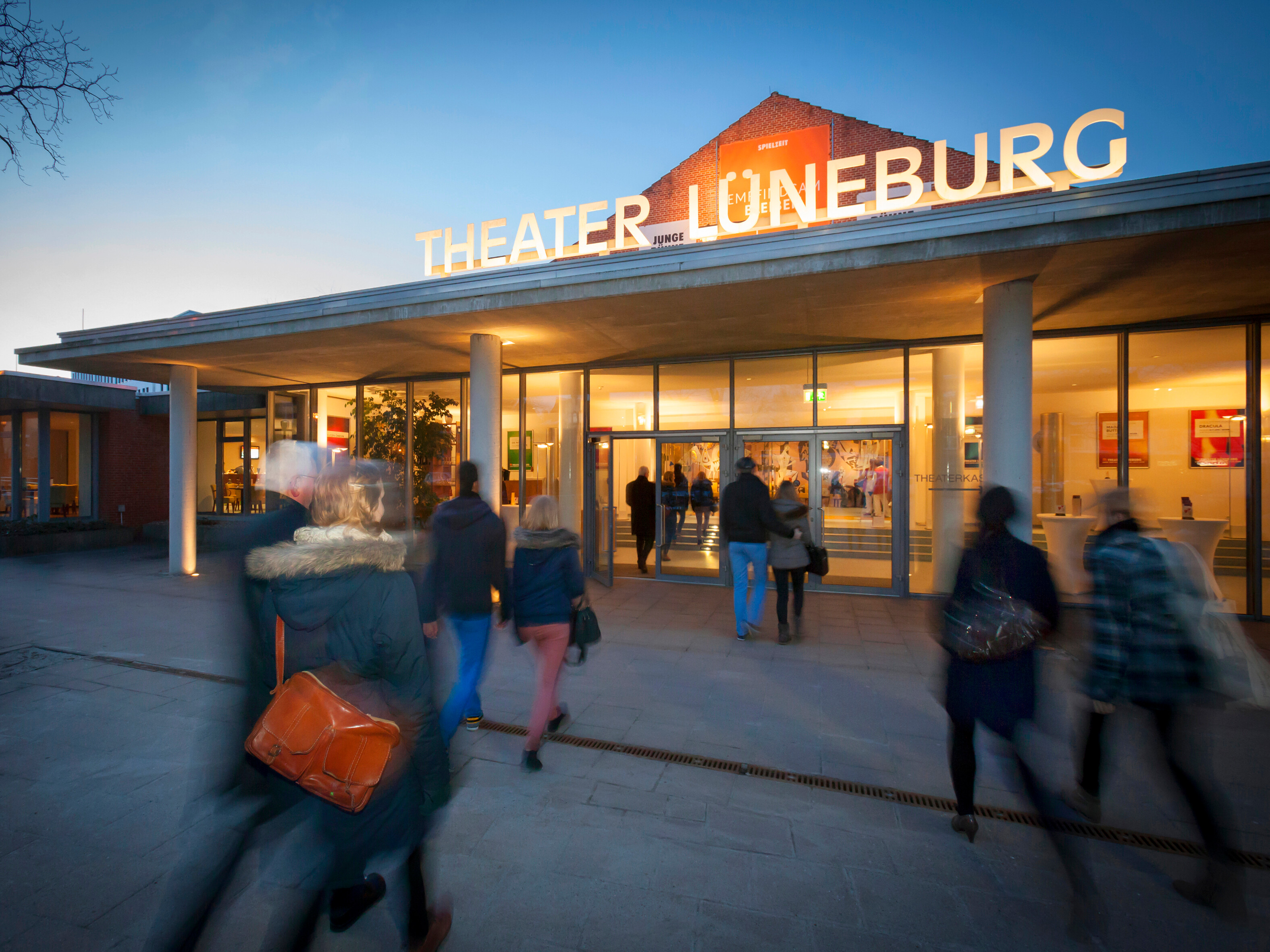 Besucher betreten das Theater Lüneburg am Abend, Außenansicht des beleuchteten Eingangsbereichs. Foto: Dan Hannen.