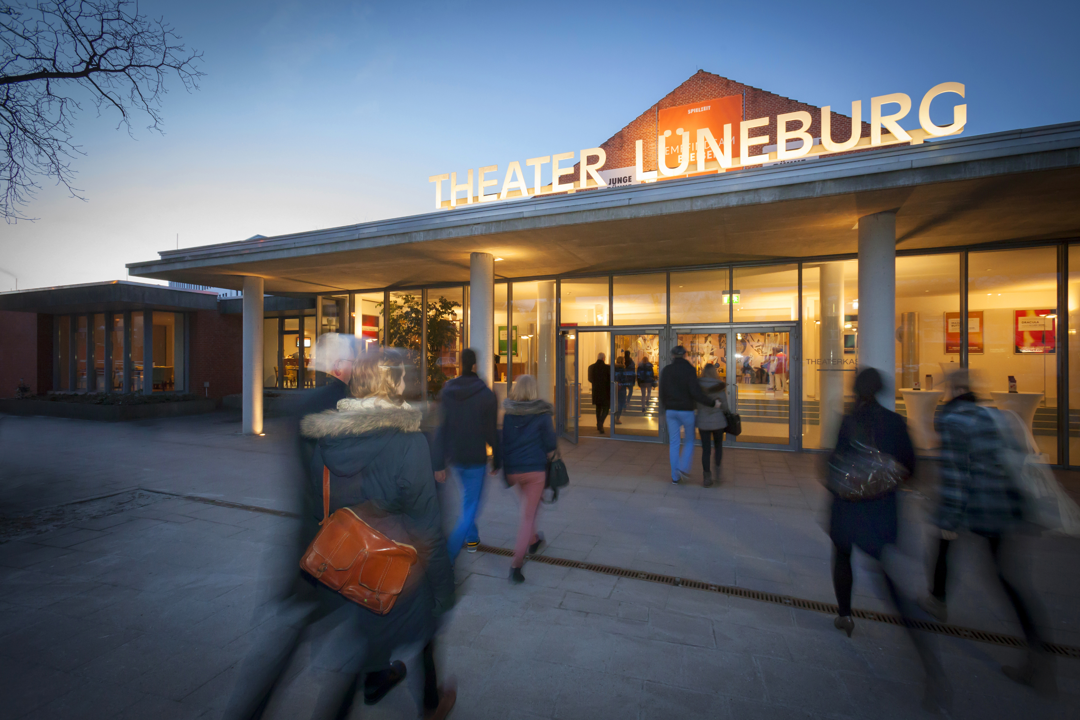 Außenansicht des Theaters Lüneburg am Abend mit Menschen, die das Gebäude betreten, beleuchteter Beschilderung und einem modernen architektonischen Design.