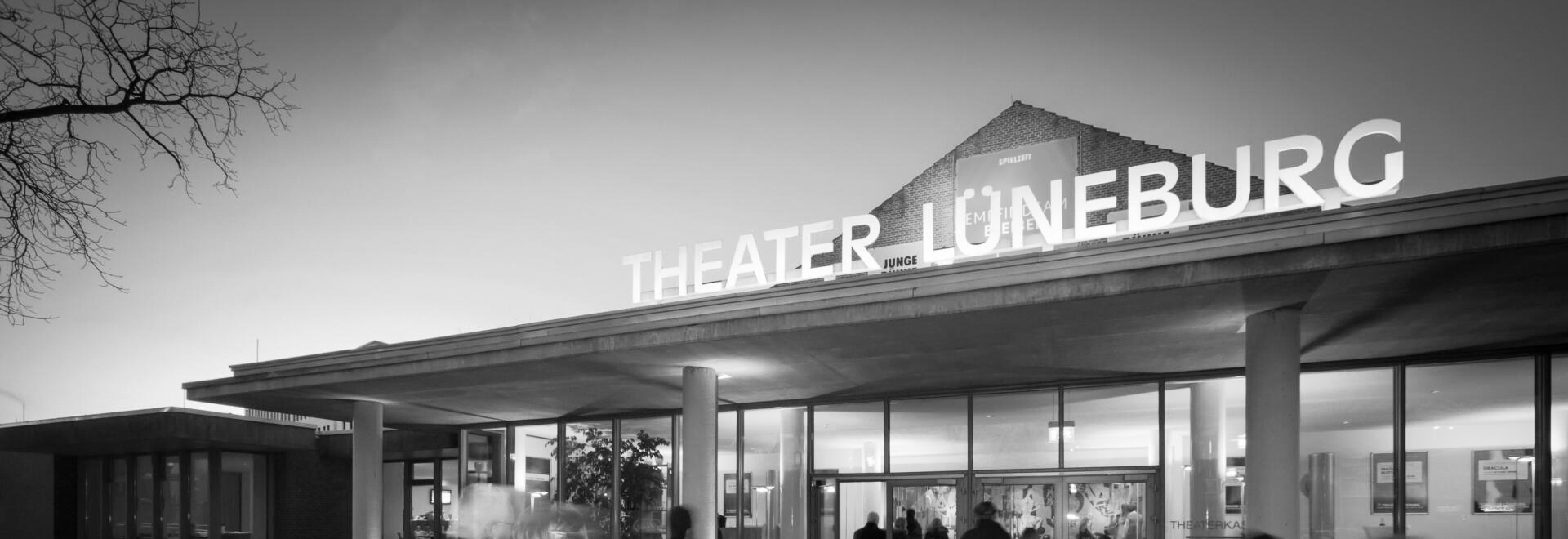 Besucher betreten das Theater Lüneburg am Abend, schwarz-weiß Aufnahme des beleuchteten Eingangsbereichs. Foto: Dan Hannen.