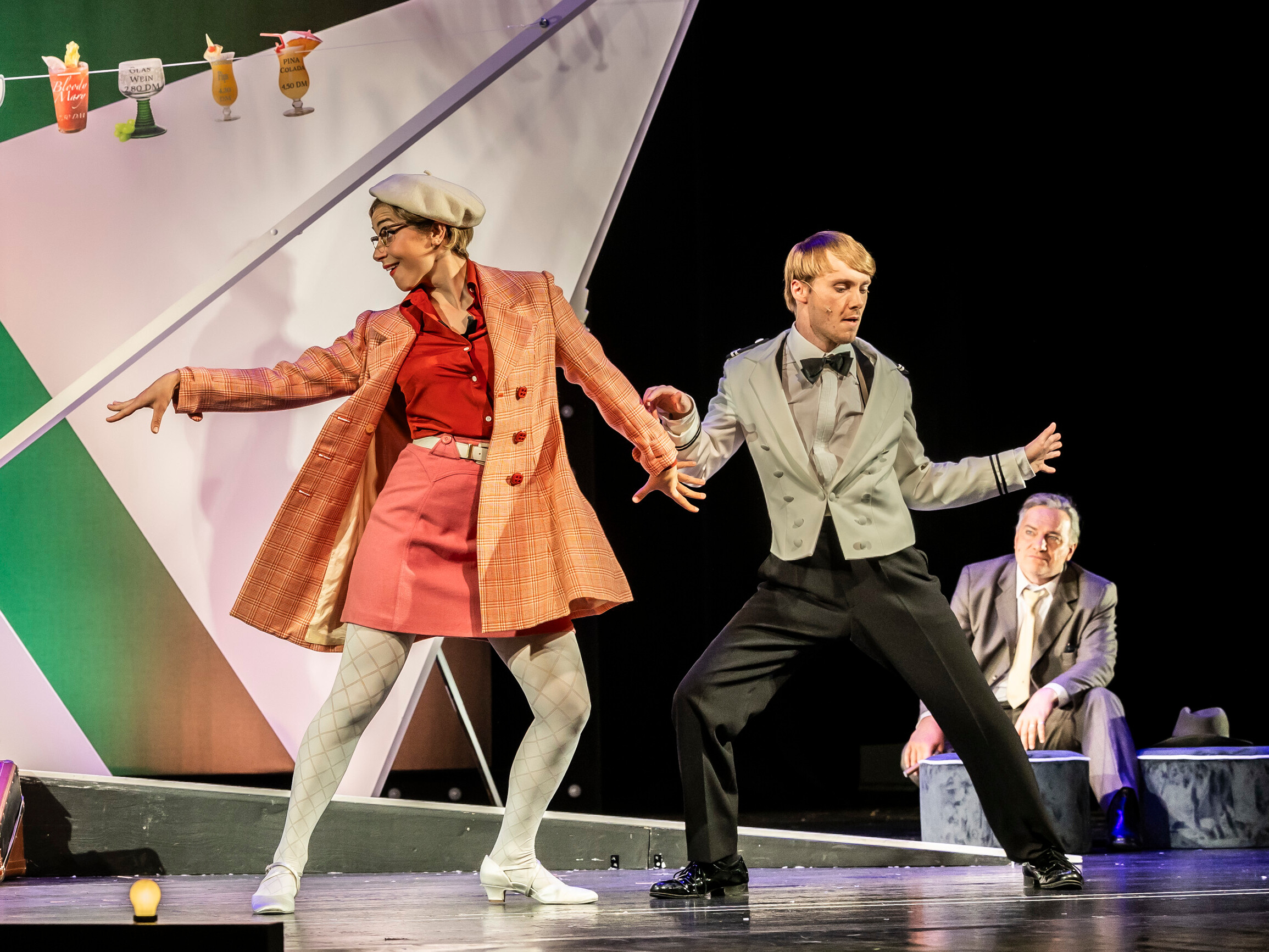 Tanzende Frau in rotem Outfit und beige Jacke, und ein Mann im grauen Smoking auf der Bühne von "Petticoat und Minirock" im Theater Lüneburg. Im Hintergrund sitzt ein Mann in einem Anzug. Foto: Jochen Quast.